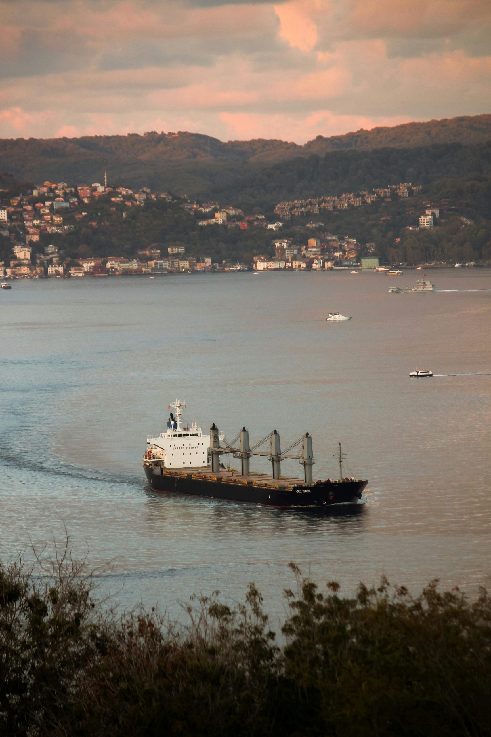 A cargo ship sails near a coastal town with hills in the background at sunset.